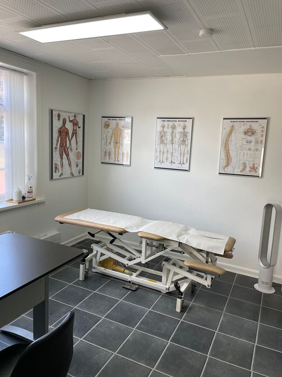 På billedet er der en briks og på vækkende hænger der 3 anatomi plakater. Der står også en fane til at køle patienten ned.